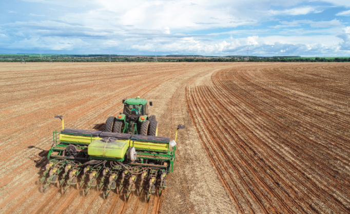 Imagem: Fotografia. Um trator verde sobre uma plantação extensa.   Fim da imagem.