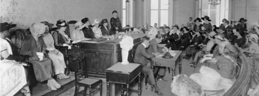 Imagem: Fotografia em preto e branco. À esquerda, homens com ternos e mulheres com vestidos estão sentados atrás de uma mesa grande. À direita, mais pessoas sentadas estão observando. Fim da imagem.