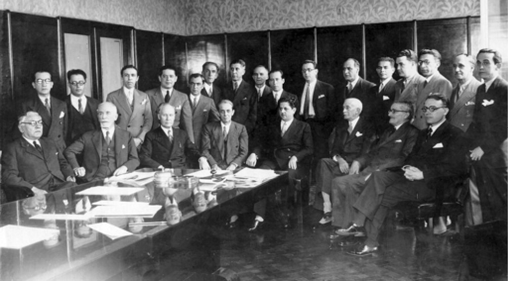 Imagem: Fotografia em preto e branco. Vários homens com terno. Alguns estão sentados e outros em pé. Na frente deles há uma mesa extensa com papéis em cima. Fim da imagem.