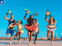 Imagem: Fotografia B. Pessoas com roupas coloridas estão segurando guarda-chuvas coloridos e pequenos e dançando.  Fim da imagem.