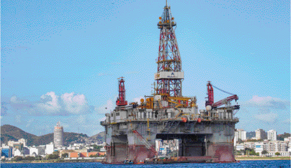 Imagem: Fotografia. Uma plataforma com uma torre alta no mar. Ao fundo, uma cidade com casas, prédios e árvores.  Fim da imagem.