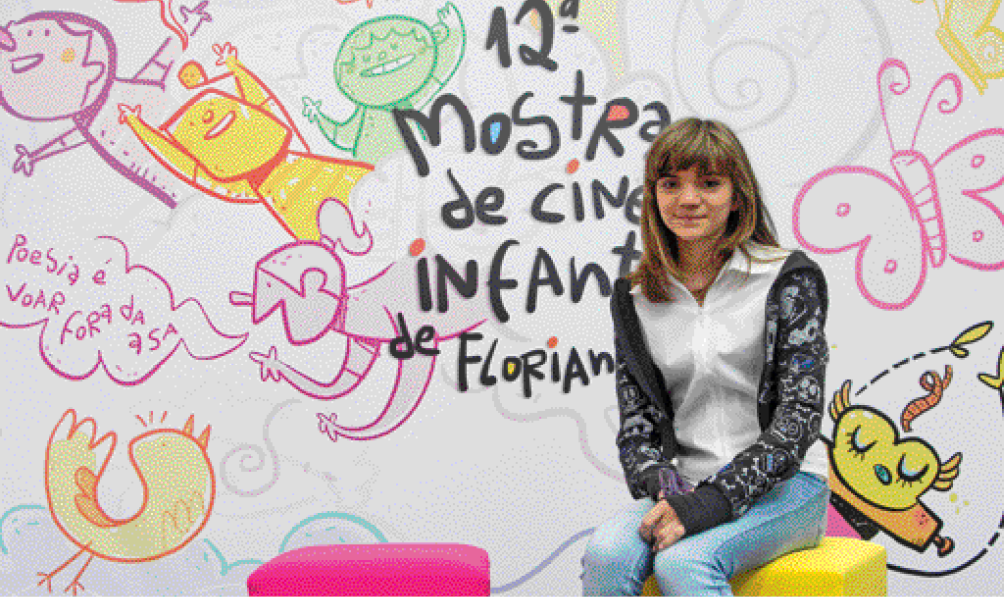 Imagem: Fotografia. Isadora Faber, menina loira com blusa preta e calça jeans está sentada e sorrindo. Atrás dela há um painel grande com desenhos e a informação: 12ª MOSTRA DE CINEMA INFANTIL DE FLORIANÓPOLIS.  Fim da imagem.