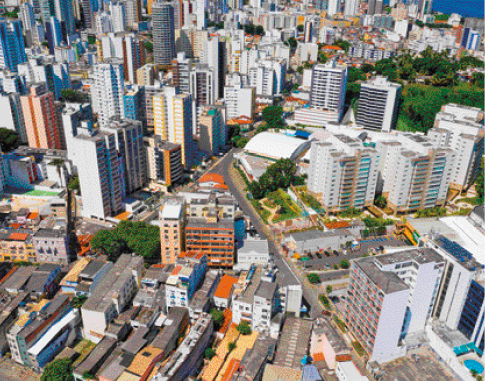 Imagem: Fotografia. Vista de cima de uma cidade com ruas, prédios, casas e árvores.   Fim da imagem.