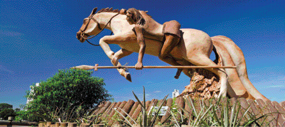 Imagem: Fotografia. Escultura de um homem com pano em volta da cintura. Ele está montado em um cavalo com o corpo inclinado para frente e segurando uma lança deitada.  Fim da imagem.