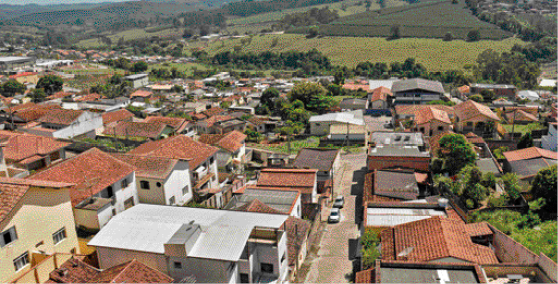 Imagem: Fotografia. Vista aérea de uma cidade com ruas, carros, casas e árvores. Ao fundo, plantações e mais árvores. Fim da imagem.