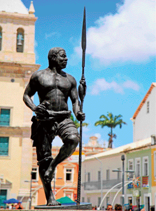 Imagem: Fotografia. Escultura de homem sem camisa com calça. Ele está segurando uma lança e com o pé esquerdo apoiado na perna direita. Ao fundo, pessoas, construções e árvores.  Fim da imagem.