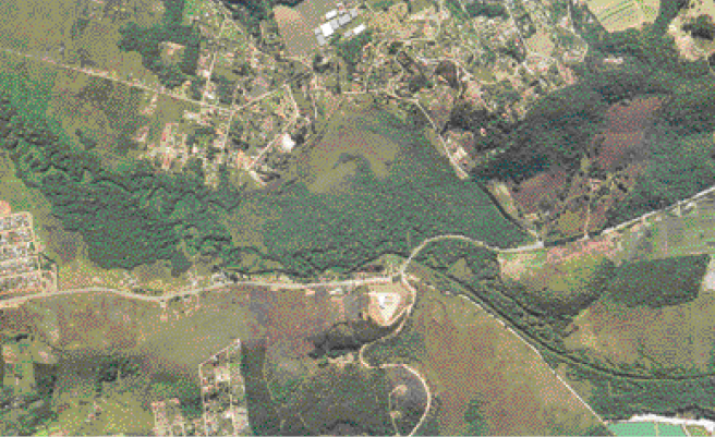 Imagem: Fotografia. Vista de cima de um município com ruas, casas, árvores e muita vegetação.  Fim da imagem.