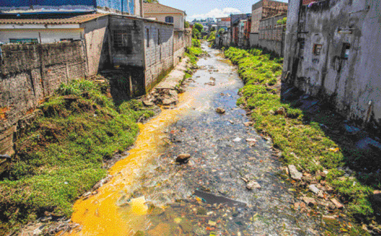 Imagem: Fotografia. Esgoto com água amarelada e muito lixo. Nas laterais há plantas e casas. Fim da imagem.