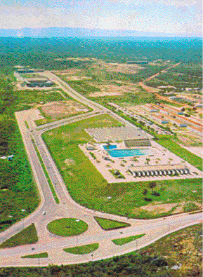 Imagem: Fotografia. Vista aérea de uma estrada asfaltada. Ao lado há uma construção grande com uma piscina e poucas construções. Ao fundo, terreno plano e vegetação.   Fim da imagem.