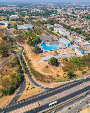 Imagem: Fotografia. Vista aérea de uma estrada asfaltada. Ao lado há uma construção grande com uma piscina e muitas construções e árvores. Ao fundo, mais construções.   Fim da imagem.