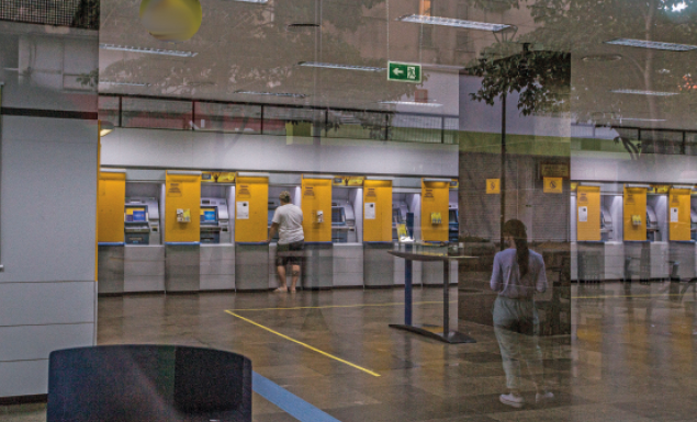 Imagem: Fotografia. Uma pessoa na frente de um caixa eletrônico. Ao lado há mais caixas eletrônicos. Fim da imagem.