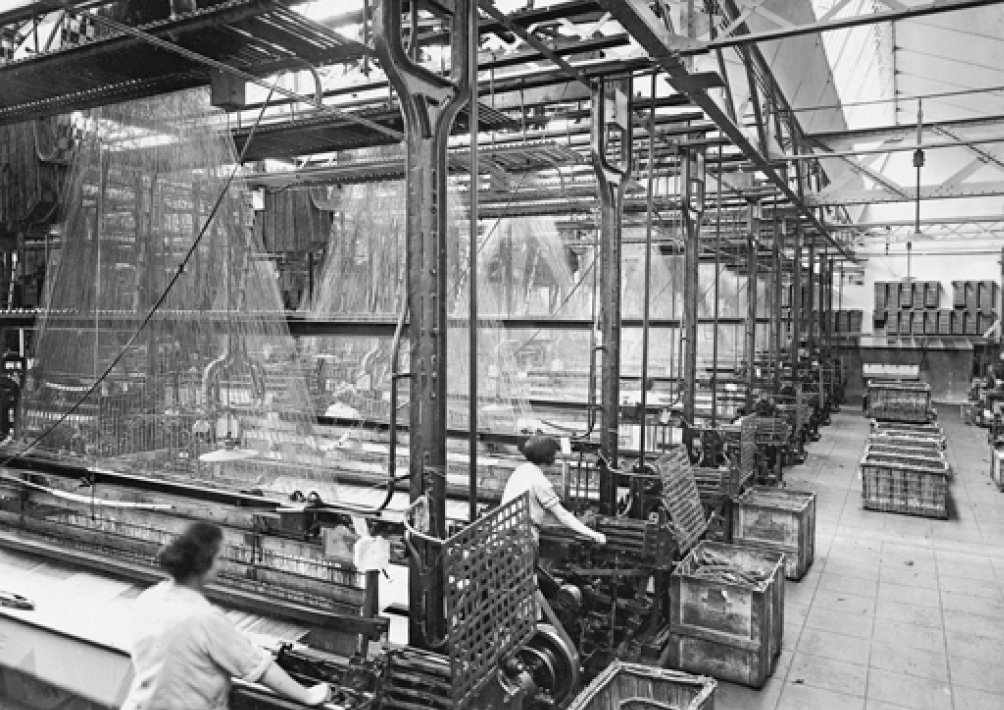 Imagem: Fotografia em preto e branco. Duas mulheres atrás de máquinas grandes com tecidos pendurados em um galpão. Fim da imagem.