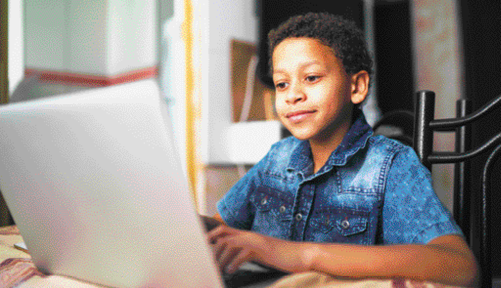 Imagem: Fotografia. Um menino com camisa azul está sentado e com as mãos no teclado de um notebook. Fim da imagem.