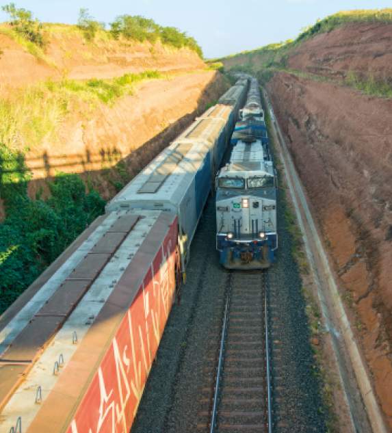 Imagem: Fotografia. Vista aérea de um trem sobre trilhos e nas laterais há morros com vegetação.   Fim da imagem.