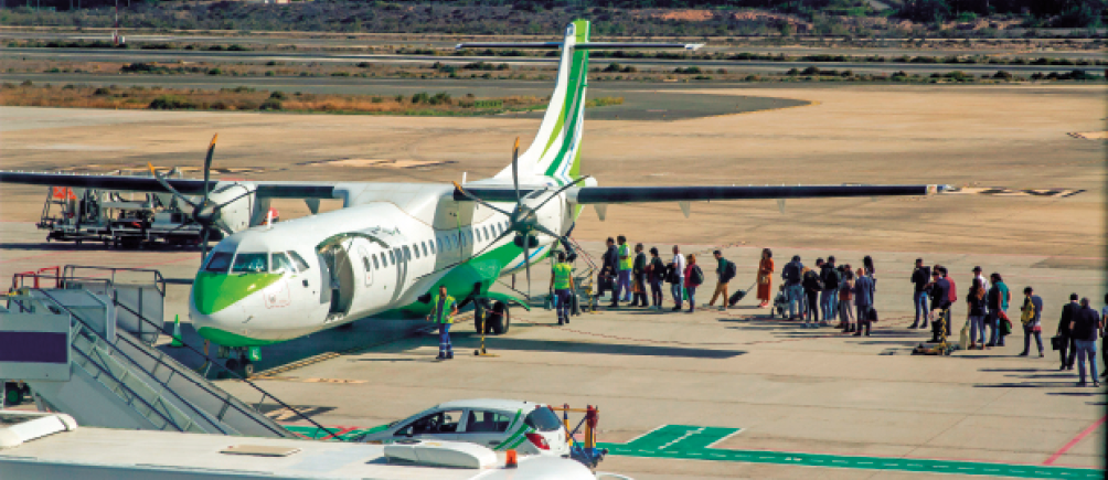 Imagem: Fotografia. Vista aérea de pessoas formando uma fila ao lado de um avião com a porta aberta.  Fim da imagem.