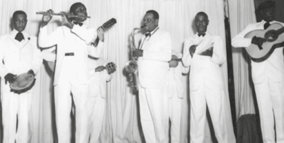 Imagem: Fotografia em preto e branco. Cinco homens com terno branco estão tocando instrumentos musicais.  Fim da imagem.