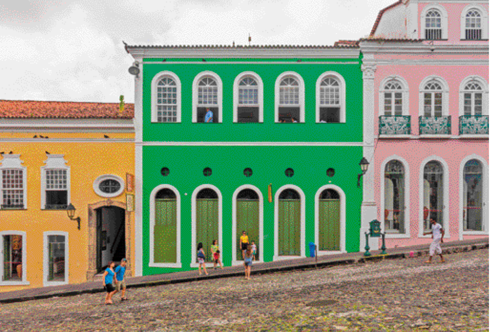 Imagem: Fotografia. Pessoas andando em uma rua íngreme de pedras. Atrás há construções lado a lado com paredes coloridas e detalhes brancos. Fim da imagem.