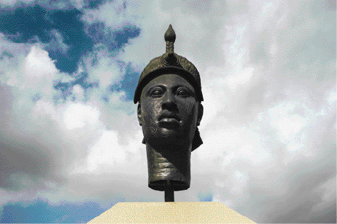 Imagem: Fotografia. Escultura da cabeça de um homem com chapéu, olhos grandes, nariz largo e lábios grossos. Fim da imagem.