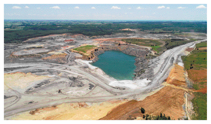 Imagem: Fotografia. Vista aérea de um lago grande com extração em volta. Ao fundo, morros e vegetação.  Fim da imagem.