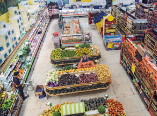 Imagem: Fotografia. Vista aérea de uma senhora em um supermercado. Ao lado dela há frutas e legumes sobre mesas. Ao redor há várias prateleiras com produtos.   Fim da imagem.