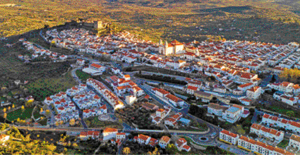 Imagem: Fotografia. Vista aérea de um castelo grande e ao redor há várias casas, ruas e árvores. Ao fundo, morros e vegetação. Fim da imagem.