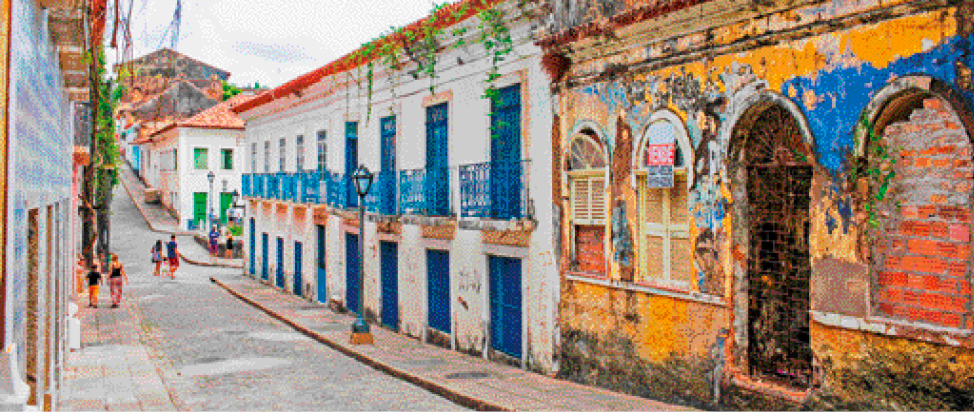 Imagem: Fotografia. Casas com paredes descascando e uma rua estreita.  Fim da imagem.