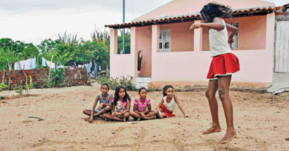 Imagem: Fotografia. Uma menina está pulando e ao seu lado, quatro meninas estão sentadas no chão e observando. Ao fundo, uma casa e plantas. Fim da imagem.