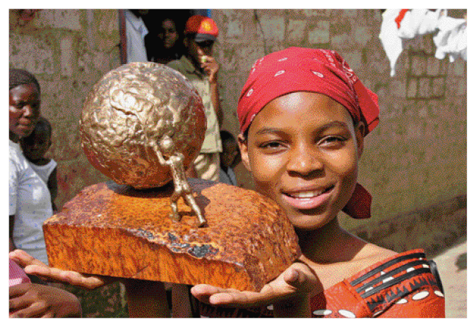 Imagem: Fotografia. Thandiwe Chama, menina negra com lenço vermelho na cabeça e vestido. Ela está sorrindo e segurando um troféu dourado com o formato de uma pessoa empurrando uma esfera grande. Ao fundo, pessoas e uma casa de barro.  Fim da imagem.