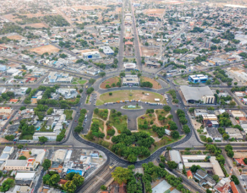 Imagem: Fotografia. Vista de cima de uma cidade com ruas, prédios, casas e árvores. No centro há uma praça com árvores.  Fim da imagem.