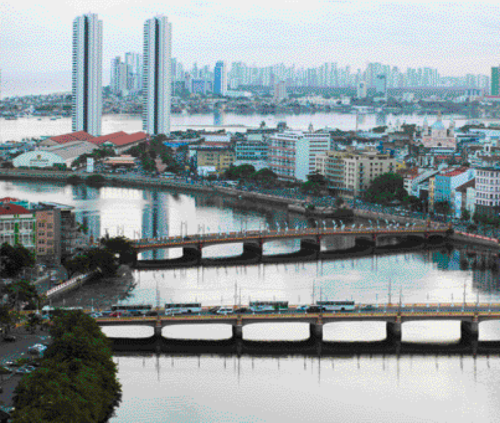 Imagem: Fotografia. No centro há duas pontes sobre um rio. Nas laterais há ruas, prédios, casas e árvores.  Fim da imagem.