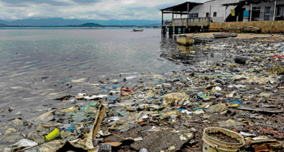 Imagem: Fotografia. Muito lixo na margem de um lago. Ao fundo, construções.   Fim da imagem.