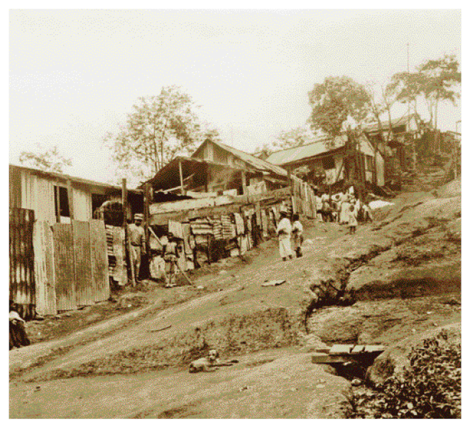 Imagem: Fotografia em sépia. Crianças em uma descida e ao lado há casas simples e um muro de telhas.  Fim da imagem.