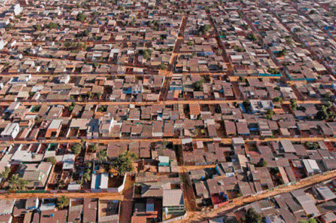 Imagem: Fotografia. Vista aérea de várias casas, árvores e ruas de terra.  Fim da imagem.