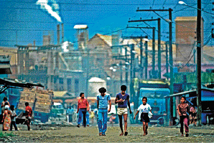 Imagem: 1980 - Cubatão foi declarada a cidade mais poluída do mundo pela Organização das Nações Unidas (ONU).  Fotografia. Crianças andando em uma rua. Ao fundo, casas, postes e chaminés de usinas expelindo fumaça.  LEGENDA: Vista da Vila Parisi, no município de Cubatão, no estado de São Paulo. Foto de 1982. FIM DA LEGENDA.    Fim da imagem.