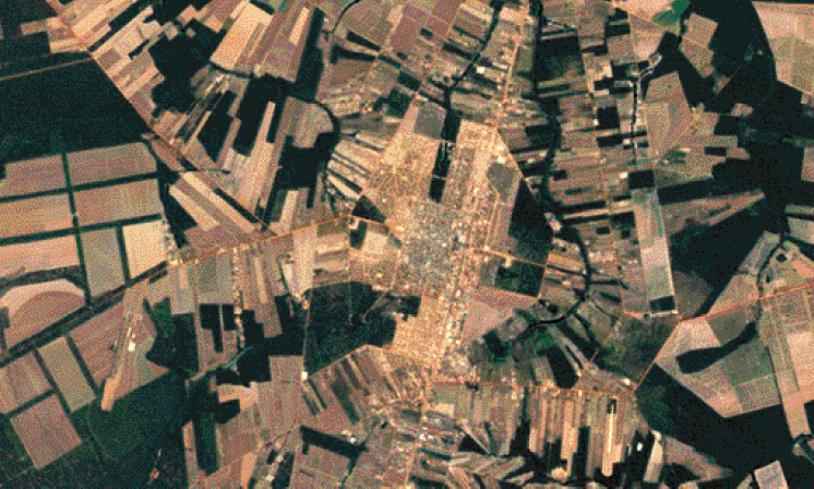 Imagem: Fotografia. Vista de cima de um município com áreas verdes e marrons. No centro há poucas construções.   Fim da imagem.