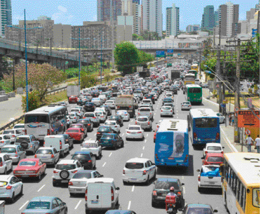 Imagem: Fotografia. Vista aérea de vários automóveis enfileirados em uma avenida. Ao fundo, prédios, árvores e casas. Fim da imagem.