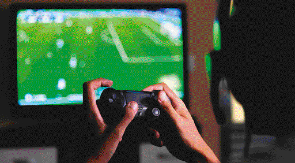 Imagem: Fotografia. Duas mãos segurando um controle de videogame. Ao fundo, uma televisão com um campo de futebol na tela. Fim da imagem.