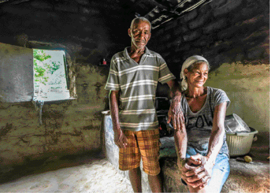 Imagem: Fotografia. Uma senhora está sentada com as mãos sobre as pernas. Ao seu lado, um senhor está em pé e com a mão sobre o ombro dela. Eles estão dentro de uma casa simples. Fim da imagem.