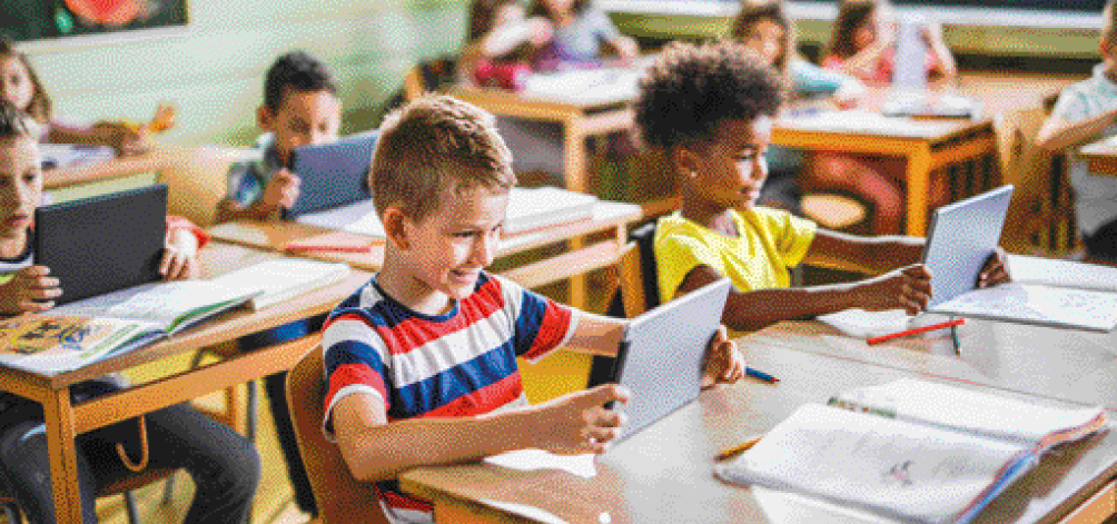 Imagem: Fotografia. Crianças sentadas em carteiras escolares e segurando tablets. Na frente delas há livros abertos.  Fim da imagem.