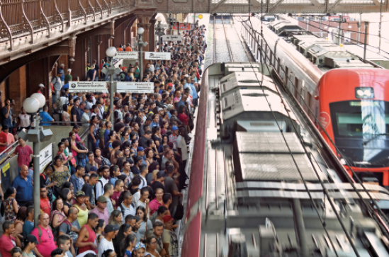 Imagem: Fotografia. Vista aérea de muitas pessoas aglomeradas na plataforma ao lado de um trem.  Fim da imagem.