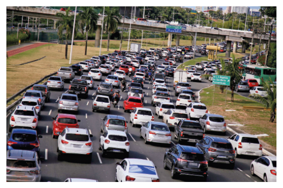 Imagem: Fotografia. Vista aérea de vários carros parados em uma avenida.  Fim da imagem.