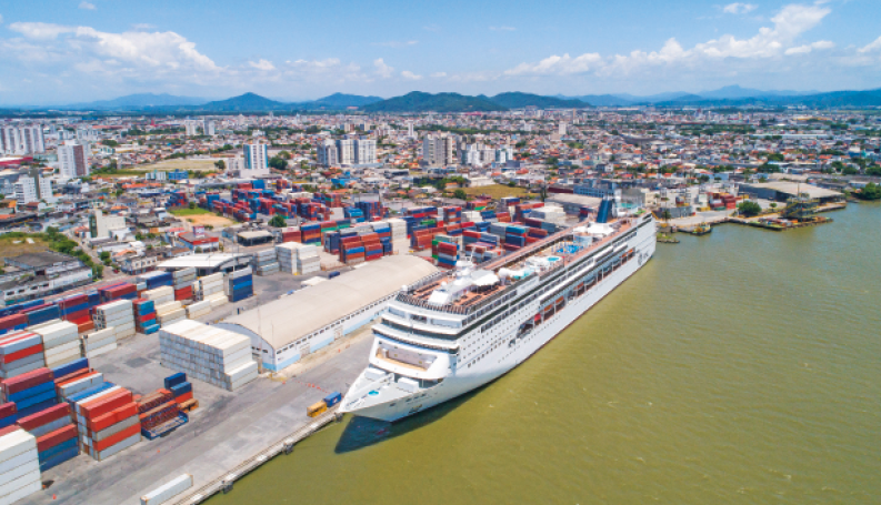 Imagem: Fotografia. Vista aérea de um navio cruzeiro gigante parado em um porto. Ao lado há milhares de contêineres empilhados. Ao fundo, uma cidade e morros.  Fim da imagem.