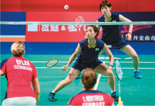 Imagem: Fotografia. Na parte superior, duas mulheres japonesas usando uniforme azul segurando raquetes. Elas olham para cima na direção de uma bola. Abaixo, uma rede e duas mulheres com cabelos loiros usando uniforme vermelho. Elas estão de costas e também seguram raquetes.  Fim da imagem.