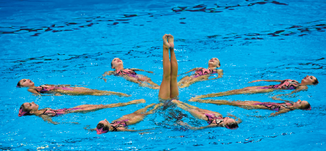Imagem: Fotografia. Oito meninas em uma piscina nadando fazendo um círculo com o corpo, no centro uma menina com as pernas esticadas para fora da água.  Fim da imagem.