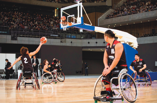 Imagem: Fotografia. Homens com uniformes pretos, sentados em cadeira de rodas em uma quadra. Um deles está com uma bola de basquete na mão. No fundo, arquibancada com pessoas.  Fim da imagem.