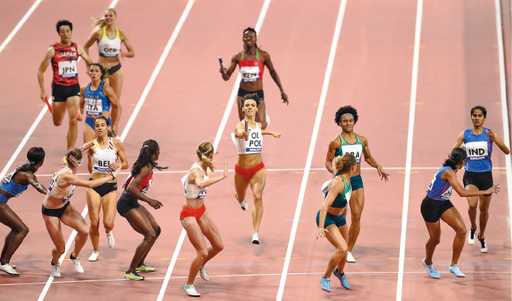 Imagem: Fotografia. Seis mulheres em uma pista de corrida olhando para trás com o braço esticado. Atrás, outras mulheres correndo segurando um bastão.  Fim da imagem.