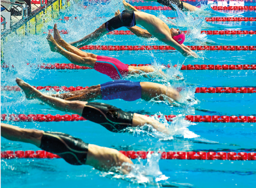 Imagem: Fotografia. Vários homens usando calção, saltando de costas na direção da piscina dividida por raias. Fim da imagem.