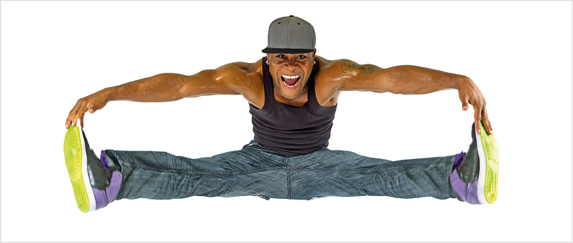 Imagem: Fotografia. Um homem negro, usando boné, regata preta, calça jeans e tênis. Ele está com as pernas na diagonal, segurando a ponta dos pés com as mãos, com o tronco flexionado para frente.  Fim da imagem.