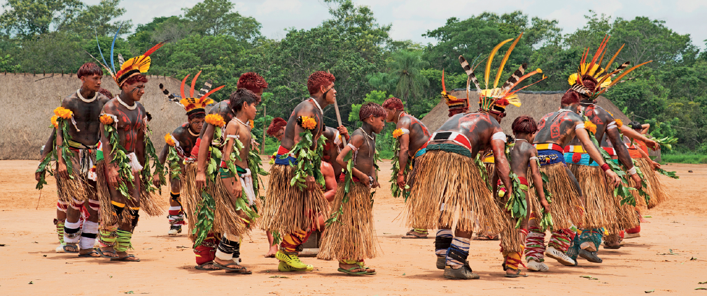 Imagem: Fotografia. Um grupo de indígenas com o corpo pintado, cocar na cabeça e saia de palha. Eles estão em um círculo com o corpo inclinado para frente. Fim da imagem.
