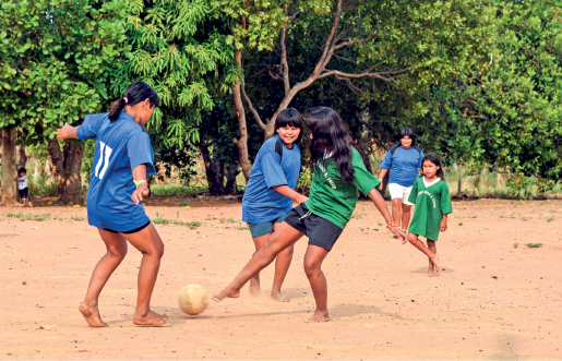 Imagem: Fotografia. Meninas, com blusas verdes e azuis chutando uma bola em um campo de terra. No fundo, há árvores.  Fim da imagem.